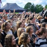 Tłum ludzi na plenerowym wydarzeniu, radosne twarze. Namiot w tle, zielone drzewa. Słoneczny dzień, atmosfera festiwalu.