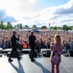 Troje wykonawców na scenie przed tłumem ludzi na festiwalu. Tło wypełnione kolorowymi namiotami, niebo z chmurami. Dominuje zielona sceneria.