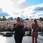 Koncert na świeżym powietrzu; tłum ludzi przed sceną. Słońce przebija się przez chmury na niebieskim niebie. Artysta z mikrofonem i kobieta na scenie.