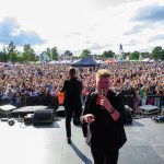 Koncert na świeżym powietrzu z dużą publicznością na tle jasno-niebieskiego nieba. Dwóch artystów w ciemnych ubraniach na scenie. W tle kolorowe namioty i atrakcje.