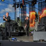 Na scenie zespół muzyczny w czarnych strojach; kobieta gra na saksofonie. Tło to jasny ekran LED i błękitne niebo.