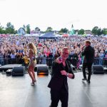 Na zdjęciu widzimy koncert na świeżym powietrzu. Muzyk z mikrofonem na scenie w czarnym ubraniu. W tle tłum ludzi i dmuchane atrakcje.
