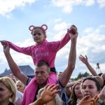 Dziewczynka w różowym ubraniu siedzi na ramionach mężczyzny w tłumie. Tło to błękitne niebo z chmurami. Wokół widać innych uczestników wydarzenia.