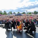 Zespół na scenie, z tłumem w tle; niebo błękitne, scenografia kolorowa. Tłoczna publika, atmosfera festiwalu.