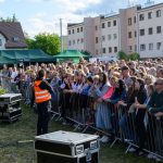 Tłum zbiera się przed sceną. Osoba w kamizelce odblaskowej stoi z przodu. W tle widać budynki i namioty.