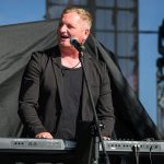 Mężczyzna w czarnej marynarce gra na keyboardzie na scenie. Jasne tło, reflektory z lewej strony. Mikrofon przed nim.