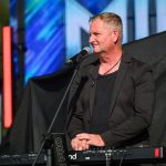 Mężczyzna w czarnym stroju stoi przy keyboardzie, trzymając mikrofon. W tle kolorowe światła sceniczne. Klasyczny koncertowy nastrój.