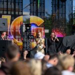 Koncert na scenie plenerowej z jasnymi światłami i kolorowym tłem. W centrum wokalistka w złotej sukni. Tłum z uniesionymi rękami na pierwszym planie.