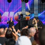 Na scenie trwa koncert; osoby z mikrofonami. Tło to kolorowy ekran LED. Przed sceną zgromadzona publiczność.
