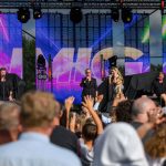 Na scenie plenerowej występują artyści, wokół nich jasne kolorowe światła. W tle widać duży ekran LED z wizualizacjami. Przed sceną zgromadzony tłum ludzi podnosi dłonie.