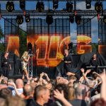 Koncert na scenie z zespołem, kolorowe światła, tłum ludzi z uniesionymi rękami. Tło to duży ekran z napisem.