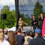 Na scenie plenerowej występują artyści wśród publiczności. Kobieta w złotej sukience gra na saksofonie, obok dwóch mężczyzn. Tło: zielone drzewa i czerwony baner.