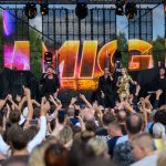 Koncert na zewnątrz z tłumem ludzi pod sceną. Jasne, kolorowe światła i ekran z napisem w tle. Cztery osoby na scenie: trzech mężczyzn i jedna kobieta.