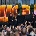 Mężczyźni w czarnych strojach śpiewają na scenie, z jasnym napisem w tle. Publiczność unosi ręce. Scena oświetlona złocistymi kolorami.