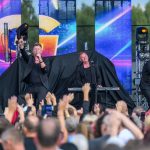 Trzech muzyków w czarnych strojach występuje na scenie. Tło to kolorowe ekrany LED z abstrakcyjnymi wzorami. Publiczność podnosi ręce w rytm muzyki.