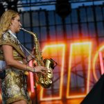 Na scenie kobieta gra na saksofonie. Ma złotą sukienkę w kwiaty, tło jest kolorowe z jasnymi światłami. Słońce odbija się na instrumencie.