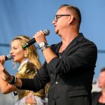Na scenie dwie osoby śpiewają z mikrofonami. Kobieta w złotej sukience i mężczyzna w czarnej marynarce. Tło jest niebieskie.