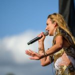 Kobieta śpiewa na scenie, trzymając mikrofon. Nosi złoto-brązowy strój z frędzlami. W tle niebo z białymi chmurami.