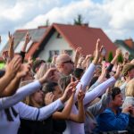 Tłum rozradowanych ludzi z podniesionymi rękoma na tle domów z czerwonymi dachami i nieba z chmurami.