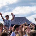 Chłopiec unosi się nad tłumem z podniesionymi rękami. Tło to niebo z chmurami i namiot. Ludzie wokół są z entuzjazmem.