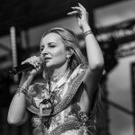 Kobieta na scenie śpiewa, trzymając mikrofon. Jest w ozdobnym stroju, z podniesioną ręką. Za nią rozmyte tło sceny.