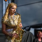Kobieta w złotej sukience gra na saksofonie na scenie. W tle ciemne i jasne elementy sceniczne. Obok niej rozmyty muzyk.