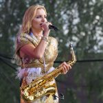 Kobieta w złotej sukni gra na saksofonie i śpiewa na scenie. Tło to rozmyte drzewa. Dominuje złoto i zieleń.