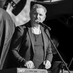 Na czarno-białym zdjęciu mężczyzna przy mikrofonie gra na klawiszach. Ubrany w ciemną marynarkę i naszyjnik. W tle ciemne zasłony sceniczne.