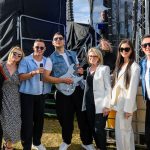 Grupa osób stoi na zewnątrz przy scenie, ubrana w kolorowe stroje, w tym jeans i biel. Na ziemi trawa, a za nimi czarne tło. Jedna osoba trzyma gitarę, a inna mikrofon.