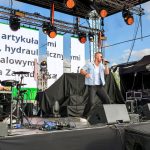 Na scenie trzech muzyków, w tle duże ekrany z tekstem i jasno-niebieskie niebo. Są światła sceniczne i sprzęt muzyczny. Artyści ubrani w jasne koszule i czarne spodnie.