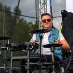Mężczyzna gra na elektronicznej perkusji na scenie. Nosi jeansową kamizelkę i ciemne okulary. Tło tworzy niebo i sprzęt sceniczny.