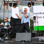 Dwóch mężczyzn na scenie, jeden śpiewa w mikrofon, drugi gra na perkusji elektronicznej. Mają na sobie jasnoniebieskie koszule. Tło to duży biały ekran i zielone akcenty świetlne.