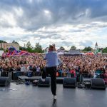 Na scenie muzycy występują przed tłumem. Niebo pochmurne, ale jasne, w tle widać kolorowe atrakcje plenerowe. Publiczność entuzjastycznie reaguje, tworząc dynamiczną atmosferę koncertu.