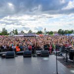 Koncert na świeżym powietrzu, z tłumem widzów pod sceną. Występujący artysta w jasnej koszuli, niebo zachmurzone. Na scenie sprzęt muzyczny, a w tle rozrywkowe budki.