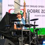 Mężczyzna w okularach przeciwsłonecznych gra na elektronicznej perkusji na scenie. W tle widoczne plakaty i reflektory. Kolory to głównie czerń, biel i zieleń.
