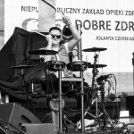 Mężczyzna gra na elektronicznej perkusji. Tło zawiera baner z napisami i sprzęt sceniczny. Zdjęcie jest czarno-białe.