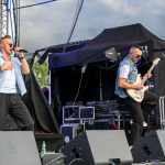 Dwóch mężczyzn występuje na scenie. Jeden śpiewa, drugi gra na gitarze elektrycznej. Tło jest ciemne z sprzętem scenicznym.