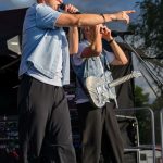 Dwóch mężczyzn śpiewa na scenie, ubranych w niebieskie kurtki. Jeden trzyma mikrofon, drugi gitarę. Tło to ciemne niebo z drzewami.