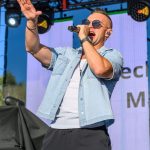 Mężczyzna śpiewa na scenie w jasnej koszuli z mikrofonem w ręku. Nosi okulary przeciwsłoneczne, a w tle widać niebieskie niebo i sprzęt sceniczny. Jest widoczne kolorowe oświetlenie sceniczne.