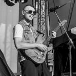 Mężczyzna gra na gitarze elektrycznej na scenie, uśmiecha się, nosi okulary przeciwsłoneczne i kamizelkę jeansową. Tło to scenografia koncertowa z oświetleniem. Zdjęcie jest czarno-białe.