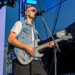 Zespół na scenie; muzyk w okularach śpiewa, trzymając połyskującą gitarę. Tło jest niebieskie, oświetlone reflektorami.