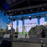 Trzech muzyków na scenie grających na instrumentach. Tło z niebieskim niebem i konstrukcją sceny. Napis TRANSPORT na ekranie.