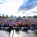 Koncert na świeżym powietrzu, widziany z perspektywy sceny. Liczna publiczność na tle nieba z chmurami. Kolorowe stoiska i zjeżdżalnia widoczne z tyłu.