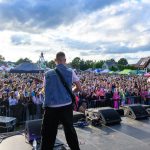 Na zdjęciu jest koncert na świeżym powietrzu z dwoma muzykami na scenie. Tłum ludzi i kolorowe stoiska w tle. Niebo jest pochmurne.