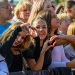 Na zdjęciu grupa uśmiechniętych osób na koncercie, pokazujących serca dłońmi. Przeważają jasne i ciepłe kolory. W tle widać tłum ludzi.