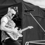 Mężczyzna gra na gitarze elektrycznej na scenie. Nosi okulary przeciwsłoneczne i dżinsową kamizelkę. Tło to ciemne kurtyny sceniczne.