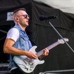 Mężczyzna gra na elektrycznej gitarze, nosi dżinsową kamizelkę. Tło sceny jest ciemne. Śpiewa do mikrofonu na statywie.