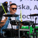 Mężczyzna w okularach gra na elektronicznej perkusji. Ma na sobie białą koszulkę i dżinsową kamizelkę. W tle widać duży ekran z tekstem.