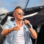 Mężczyzna w okularach przeciwsłonecznych śpiewa na scenie. Ma jasną koszulę, trzyma mikrofon. Tło jest ciemne, koncertowe.
