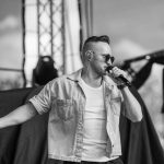 Mężczyzna w okularach przeciwsłonecznych śpiewa na scenie, trzyma mikrofon. Na sobie ma dżinsową koszulę. W tle rozmyte elementy sceny.